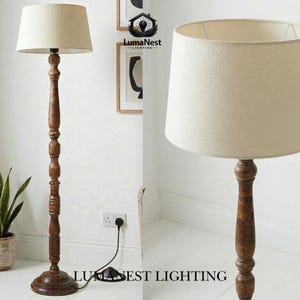 May include: A wooden floor lamp with a beige lampshade. The lamp has a dark brown, ornate base and a tall, carved wooden pole. The lampshade is a neutral color, and the lamp is plugged into a wall outlet. The text "LumaNest Lighting" is visible.