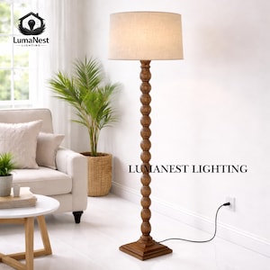 May include: A wooden floor lamp with a beige fabric shade. The lamp has a turned wood base and a series of decorative wooden spheres on the central column. The base is square. The text "LumaNest Lighting" is printed on the lamp.