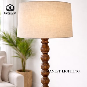 May include: A floor lamp with a beige linen shade and a dark brown, turned wood base. The lamp has a classic design and is suitable for living rooms or bedrooms. The text "LumaNest Lighting" is visible on the base.