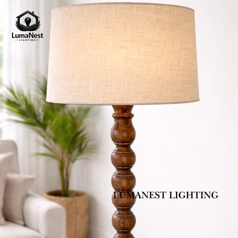 May include: A floor lamp with a beige linen shade and a dark brown, turned wood base. The lamp has a classic design and is suitable for living rooms or bedrooms. The text "LumaNest Lighting" is visible on the base.