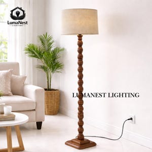 May include: A wooden floor lamp with a beige lampshade. The lamp has a dark brown, turned wood base and a matching stem. The text "LUMANEST LIGHTING" is printed on the wall. The lamp is plugged into a wall outlet.