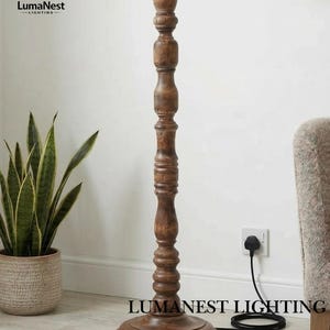 May include: A wooden floor lamp with a dark brown finish. The lamp has a turned wood design and a round base. The brand name "LumaNest Lighting" is printed on the base. A black power cord is connected to a wall outlet.