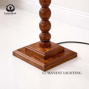 May include: A wooden floor lamp base with a square base and a turned, baluster-style column. The lamp is made of dark brown wood and has a black power cord. The base is square and has a layered design.