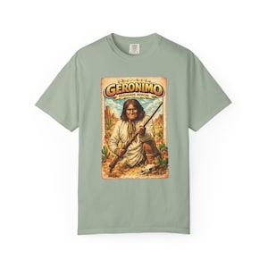Op de afbeelding: Saliegroen t-shirt met een retro graphic van Geronimo die een geweer vasthoudt in een woestijnlandschap. De graphic bevat de tekst "GERONIMO" en "NOENDAHE APACHE" in een vintage lettertype.