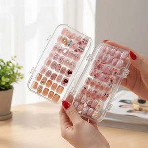Peut inclure: Deux boîtes en plastique transparent remplies de faux ongles de différentes couleurs : rose, nude et or. Certains ongles sont décorés de strass et de motifs floraux. Les boîtes sont ouvertes, révélant la variété des décorations.