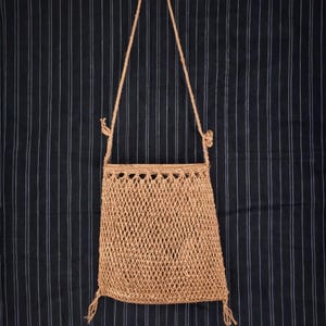 May include: A tan, woven shoulder bag with a diamond-patterned body and a braided strap. The bag has a rectangular shape with a decorative top edge and tassels at the bottom. The background is a dark blue fabric with vertical white stripes.