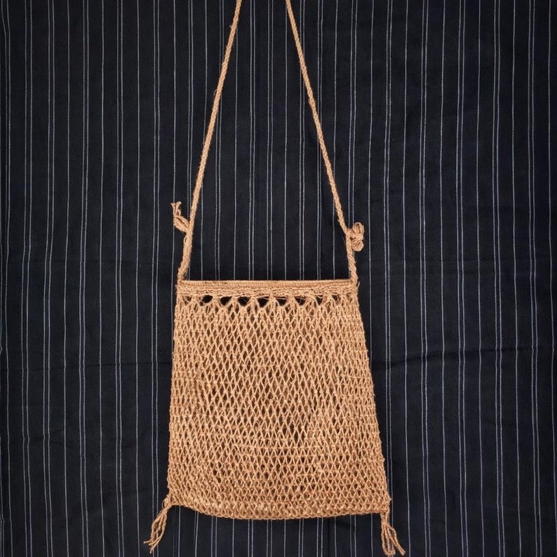 May include: A tan, woven shoulder bag with a diamond-patterned body and a braided strap. The bag has a rectangular shape with a decorative top edge and tassels at the bottom. The background is a dark blue fabric with vertical white stripes.