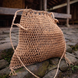 May include: A woven, open-weave bag in a natural brown color. The bag has a rounded bottom and a drawstring closure at the top. The straps are made of the same material as the bag. The bag is sitting on a stone surface.