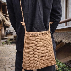 May include: A tan, open-weave shoulder bag with a long strap. The bag is made of a woven material, with a rectangular shape and a tassel at the bottom. The bag is worn over the shoulder.
