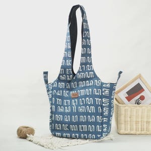 May include: A blue tote bag featuring a repeating white abstract pattern and a small brown rectangular brand label on the front. The bag has two long shoulder straps and side extensions, with a structured design and a solid dark interior lining.