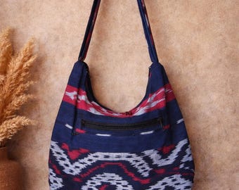 May include: A blue shoulder bag with a patterned design in red and white. The bag has a black zipper and a fringe at the bottom. The bag is hanging against a neutral background.
