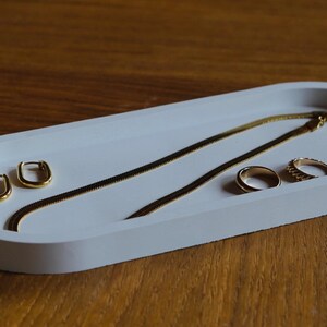 Può includere: Un vassoio portagioie bianco di forma ovale con orecchini, collane e anelli dorati. Il vassoio è realizzato in materiale liscio e poggia su una superficie in legno. I gioielli includono una collana a catena d'oro e due anelli.