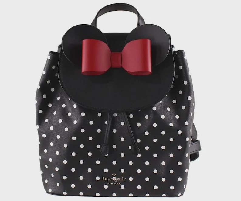 May include: Black and white polka dot backpack with a red bow and mouse ears. The backpack has a top handle and adjustable straps. The brand name "kate spade new york" is visible on the front.