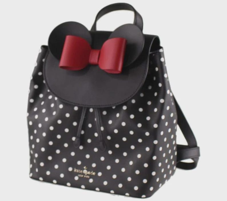 May include: Black backpack with white polka dots, featuring a black flap with a red bow and mouse ears. The bag has a drawstring closure and a top handle. The brand name "kate spade new york" is visible on the front.