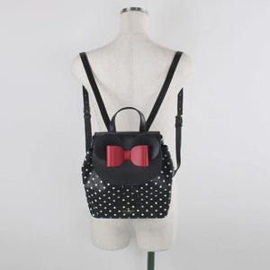 May include: Black backpack with white polka dots and a red bow. The backpack has adjustable black straps and a top handle. The flap features a red bow and the shape of mouse ears. The bag is displayed on a mannequin.