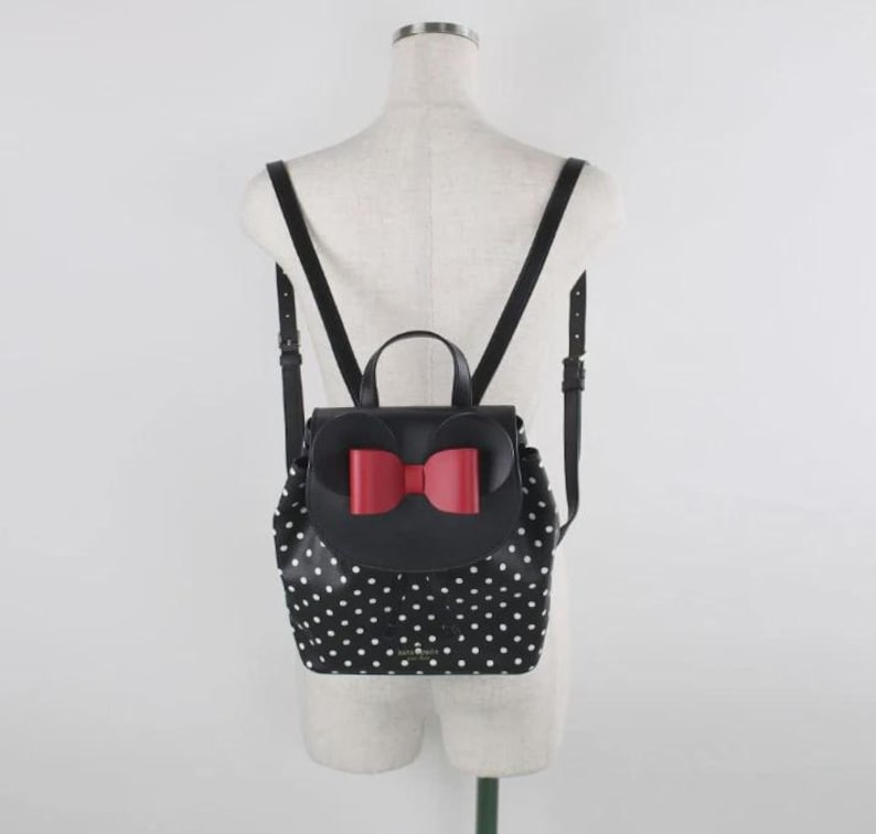 May include: Black backpack with white polka dots and a red bow. The backpack has adjustable black straps and a top handle. The flap features a red bow and the shape of mouse ears. The bag is displayed on a mannequin.