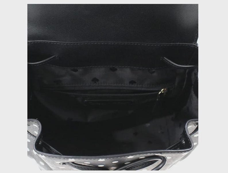 May include: Black backpack with white polka dots, open to reveal the interior. The interior is black with a small zippered pocket and a lining with a repeating spade pattern. The bag has a drawstring closure and a flap.