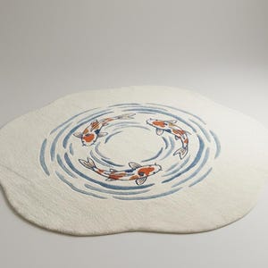 May include: A cream-colored, oval rug featuring a design of three koi fish swimming in a circle. The fish are orange, white, and black, with blue water lines. The rug has a soft, textured appearance.
