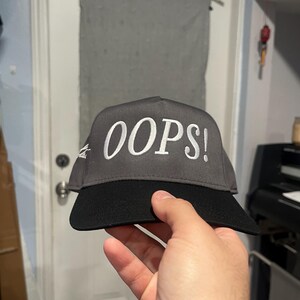 Peut inclure: Casquette de baseball grise et noire avec le mot "OOPS!" brodé en blanc. La casquette a un design structuré avec une visière incurvée. Le chapeau est tenu par une main.