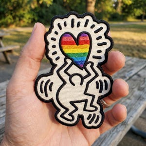 May include: Embroidered patch featuring a stylized figure holding a rainbow heart. The patch has a cream-colored background with black outline and details. The heart displays the colors of the rainbow.