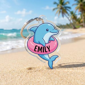 May include: A clear acrylic keychain featuring a cartoon dolphin in a pink inner tube. The dolphin is light blue with a smiling face. The name "EMILY" is printed in black on the inner tube. A silver metal keyring is attached.