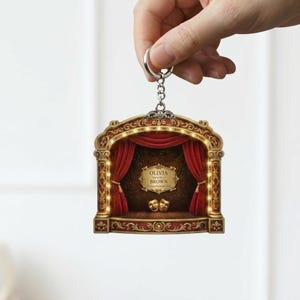 May include: A theater-themed keychain with a decorative stage design. The keychain features a red curtain, a gold-colored frame, and the text "OLIVIA BROWN". The stage has two golden masks and is illuminated with small lights.