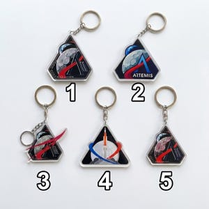 May include: Five keychains with space-themed designs. Each keychain features a triangular emblem with a silver keyring. The designs include rockets, planets, and the word "ARTEMIS". The keychains are numbered 1 through 5.