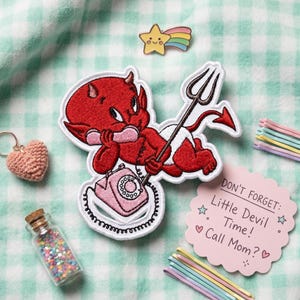 May include: An embroidered patch featuring a red devil character holding a black trident and talking on a pink rotary telephone. The patch is surrounded by a small star pin, a crochet heart keychain, a glass bottle of colorful beads, and a pink note.
