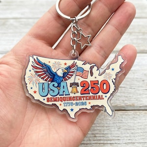 May include: A clear acrylic keychain shaped like the United States, featuring a patriotic design. The keychain includes a silver keyring, a star charm, and a design with an eagle, flag, and the text "USA 250 SEMIQUICENTENNIAL 1776-2026".