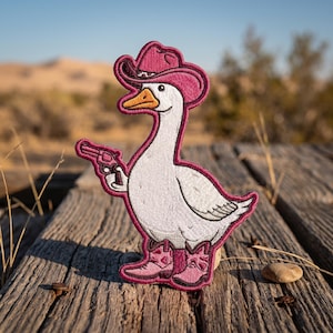 May include: An embroidered patch featuring a white goose wearing a pink cowboy hat and matching pink cowboy boots. The goose is holding a small pink revolver in its wing. The design is outlined in a dark pink border against a white background.