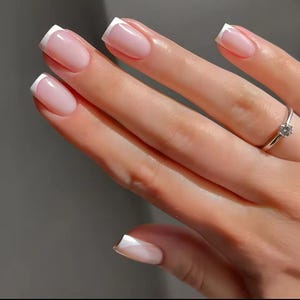 May include: Close-up of a hand with French manicure nails. The nails are a natural pink with white tips. A silver ring with a small, clear stone is on the ring finger. The background is a soft, neutral colour.