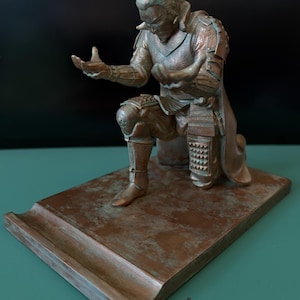May include: A bronze-coloured statue of a kneeling samurai warrior with a topknot, wearing detailed armour. The figure is positioned on a rectangular base with a grooved slot in the front, designed to hold a mobile device or similar object.