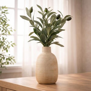 May include: A beige ceramic vase holding an olive branch with green leaves and dark olives. The vase has a rounded shape and sits on a wooden surface. The background includes a window and sheer curtains.