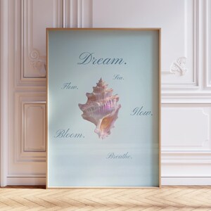 May include: A framed vertical wall art print featuring a central iridescent seashell in shades of pink and white against a pale blue background. The text Dream, Sea, Flow, Glow, Bloom, and Breathe is printed in a dark blue script font around the shell.