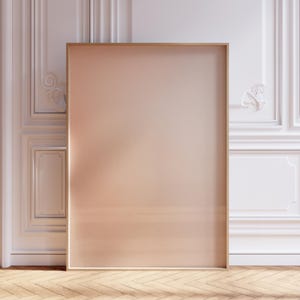 May include: A large, rectangular framed artwork with a soft peach-colored center. The frame is a light wood tone. The artwork leans against a white wall with decorative molding and sits on a light wood floor.