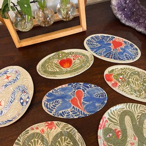 May include: A selection of oval ceramic dishes with hand-painted designs. The dishes showcase diverse motifs, such as snakes, hearts, and floral patterns in blue, red, green, and beige. The dishes are presented on a dark wooden surface.