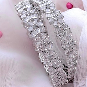 May include: Two silver bangles featuring detailed floral and geometric patterns. The bangles are embellished with many clear, sparkling stones, producing a dazzling effect. The jewellery is presented on a soft, white fabric.