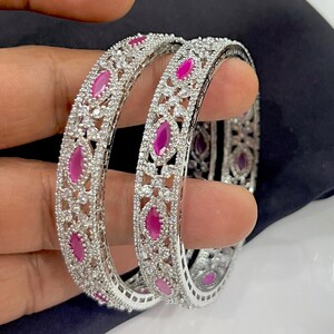 May include: Two silver-toned bangles featuring a repeating pattern of marquise-cut fuchsia pink stones alternating with clusters of small, clear, round-cut white stones set in a metallic frame with a detailed, openwork filigree design.