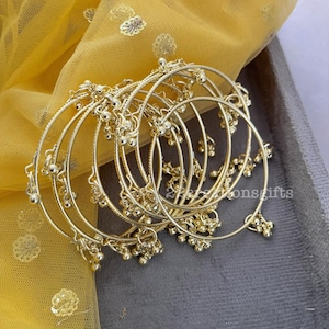 May include: A set of gold-colored bangles with small dangling beads. The bangles are arranged on a gray surface, with a yellow, sheer fabric with gold embellishments in the background. The bangles have a delicate, ornate design.