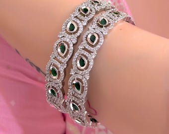 May include: Two silver bangles with a repeating pattern of teardrop-shaped emerald green stones and clear crystals. The bangles are worn on a wrist, set against a pink background.