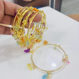Op de afbeelding: Goudkleurige armbanden met kleurrijke edelsteenaccenten. De armbanden zijn versierd met een verscheidenheid aan pastelkleurige edelstenen, waaronder roze, blauw, geel en oranje. Het ontwerp heeft een delicaat, bungelend charme-element.