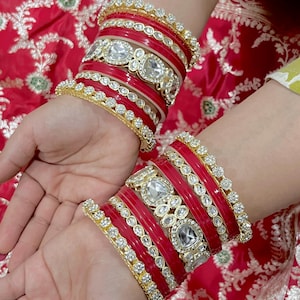 May include: A collection of red and gold bangles, adorned with clear, sparkling stones. The bangles are stacked on the wrist, displaying the detailed design and vivid colours. The jewellery is likely for a special event.