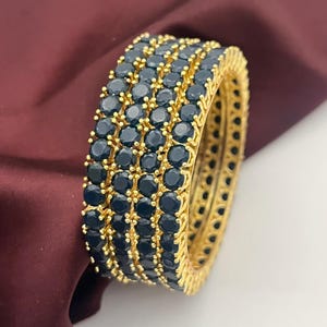 May include: A collection of gold-coloured bangles, each adorned with rows of dark blue gemstones. The bangles are stacked, highlighting the detailed design and the contrast between the gold and the dark blue stones. The background is a deep burgundy colour.
