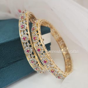 May include: Two gold-colored bangles with floral patterns. Each bangle is adorned with clear crystals and small red gemstones. The bangles are resting on a teal surface with a white fabric backdrop. The text "2creationsgifts" is visible.