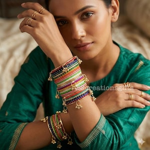 May include: A woman wearing multiple gold rings and a green top, showcasing colorful bangles. The bangles are a mix of vibrant colors, adorned with small gold bells. The image is well-lit, highlighting the jewelry's details.