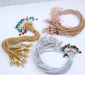 May include: A selection of decorative tassels in different colours. The tassels have gold, silver, and tan cords, decorated with red, green, and white beads. The tassels are arranged in a circular pattern against a white background.