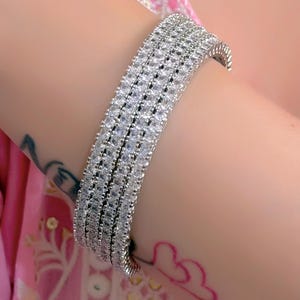 May include: A silver-toned bracelet with multiple rows of clear, sparkling stones. The bracelet is curved to fit the wrist and reflects light, creating a brilliant display. The jewelry is set against a soft pink background.