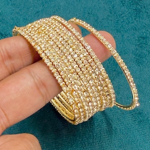 May include: A set of gold-toned bangles featuring a continuous row of small, clear, sparkling rhinestones embedded along the outer surface of each circular band. The bracelets are thin, metallic, and designed to be worn stacked together on the wrist.