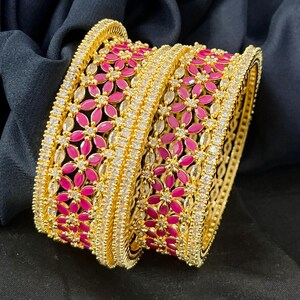 May include: A pair of gold-tone bangles featuring a central band of vibrant pink flower-shaped stones. Each bangle is bordered by two rows of clear, sparkling white stones set in a metallic frame with a textured, beaded edge design.
