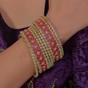 Könnte beinhalten: Ein Satz goldfarbener Armreifen, verziert mit rosa Blumenmustern und klaren, funkelnden Steinen. Die Armreifen sind an einem Handgelenk gestapelt und zeigen die komplizierten Details des Schmucks. Der Hintergrund ist ein tiefvioletter Stoff.
